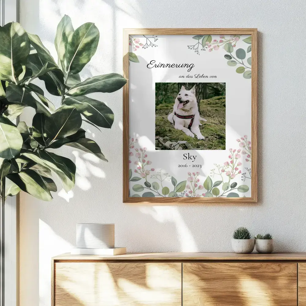 Gedenkposter mit weißem Hund, Namen und Lebensdaten im Blumenrand, im Holzrahmen über einem Sideboard