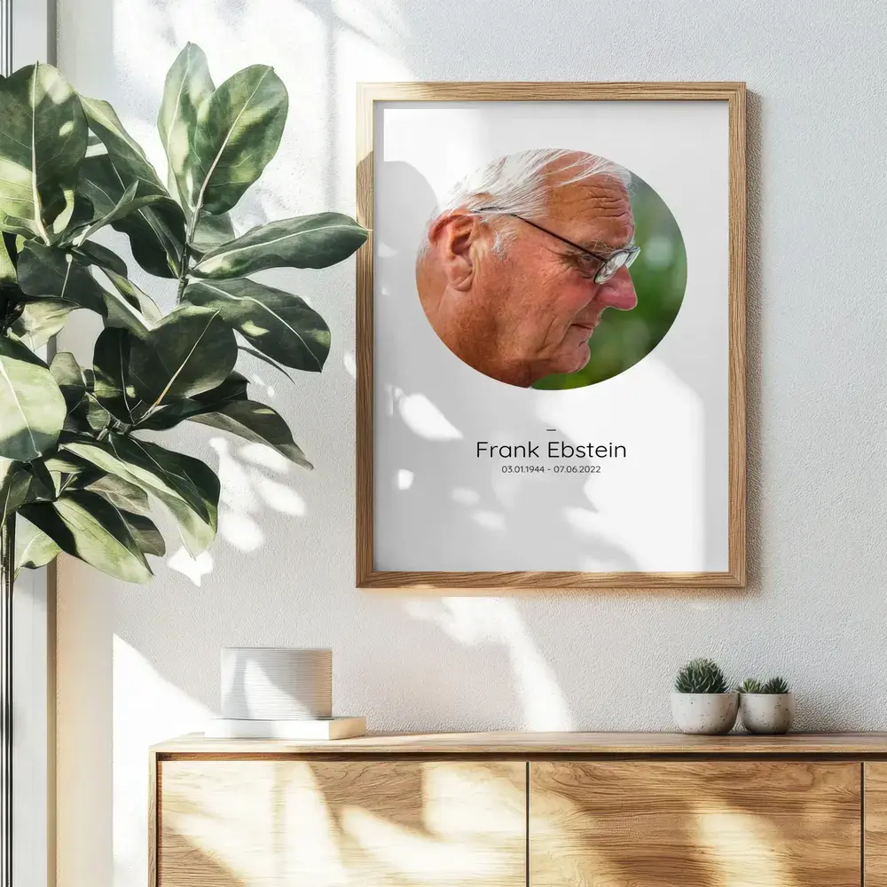 Trauerbild mit rundem Porträt und Namen im Naturholzrahmen an der Wand über einem Sideboard neben einer Pflanze