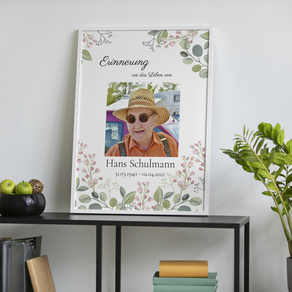 Trauerposter mit Porträt und zarten Blumen im weißen Rahmen auf einer schwarzen Konsole neben einer Pflanze