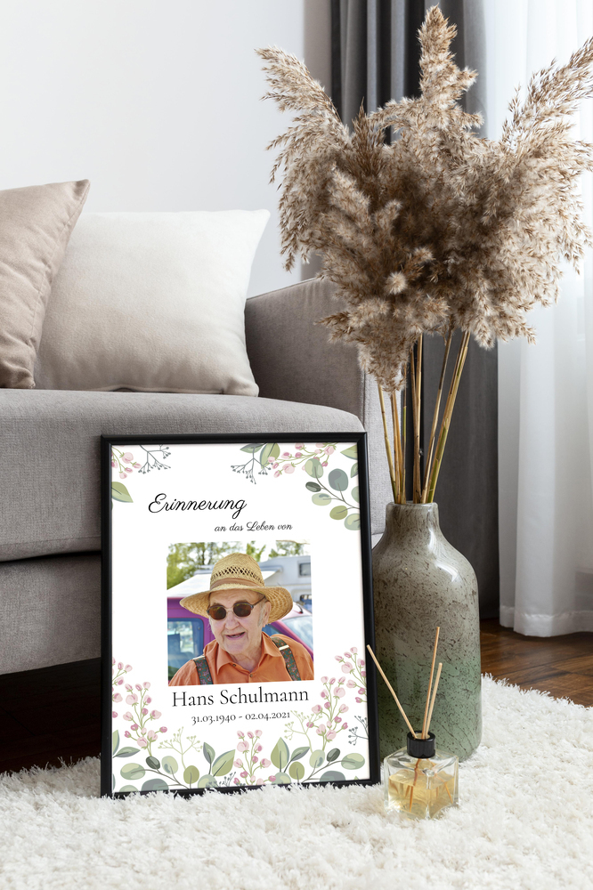 Trauerposter mit Blumenmotiv im schwarzen Rahmen auf einem hellen Teppich neben Sofa und Vase mit Trockenblumen